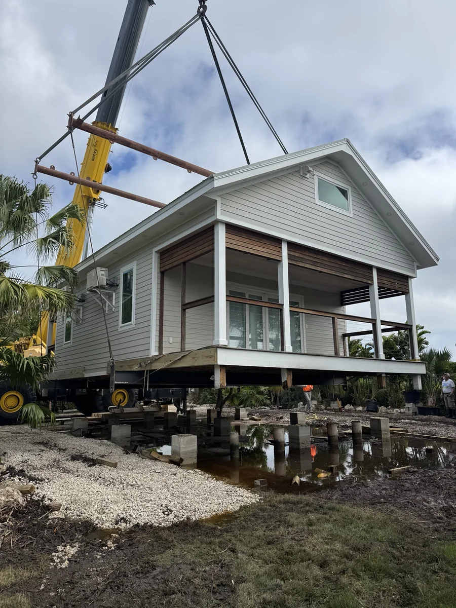 House being positioned over the prepared site with crane rigging and temporary supports.